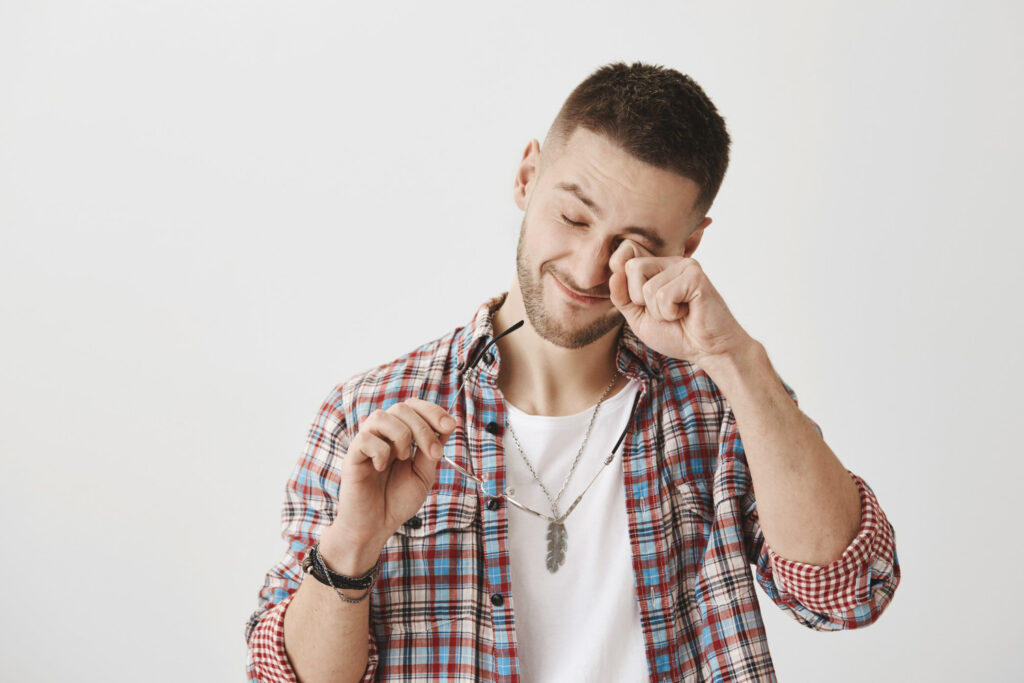 Young man holding prescription glasses on one hand and rubbing his eye with the other hand
