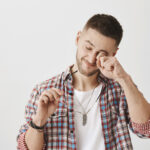Young man holding prescription glasses on one hand and rubbing his eye with the other hand